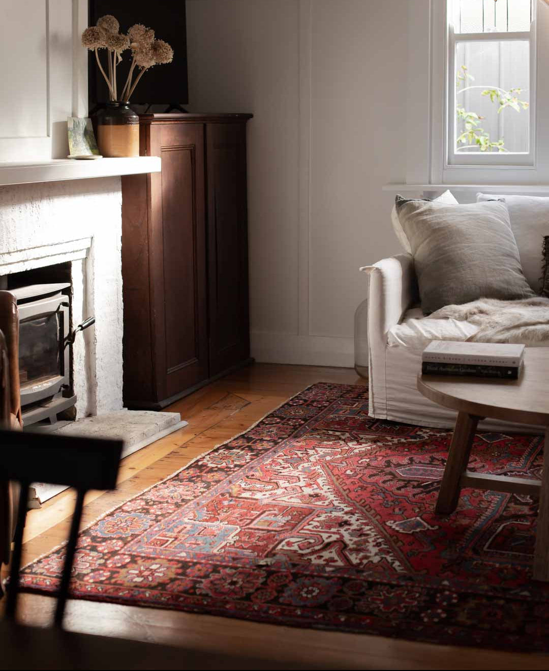A cozy living room corner featuring a traditional red and blue patterned oriental area rug on a hardwood floor, next to a white sofa with neutral pillows, a dark wood cabinet, and a white fireplace.