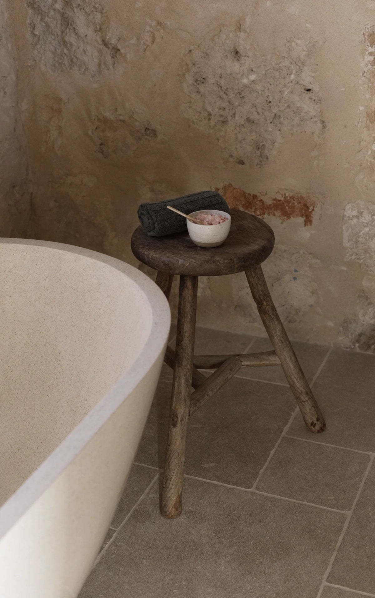 Deep stone bath with wooden stool holding bath salts and a towel